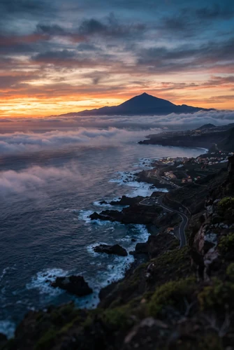 Uma vista aérea de um litoral acidentado no crepúsculo. O oceano se agita contra a costa escura e rochosa enquanto uma estrada sinuosa atravessa os povoados abaixo, e um manto de nuvens baixas se estende para o mar diante da silhueta de uma montanha distante, sob um céu com faixas laranjas e azuis.