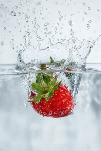 Um morango vermelho vibrante mergulha em águas cristalinas, criando um respingo dinâmico com gotas suspensas. A captura em alta velocidade congela o movimento, destacando a textura da fruta e a natureza efêmera da água. A iluminação é brilhante e limpa, enfatizando o objeto contra um fundo suavemente desfocado.